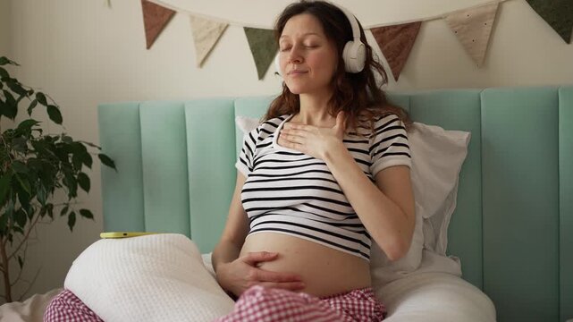Peaceful pregnant woman meditating in her bedroom, listening to calming music on headphones while gently caressing her baby bump, promoting relaxation and maternal bonding during expectancy