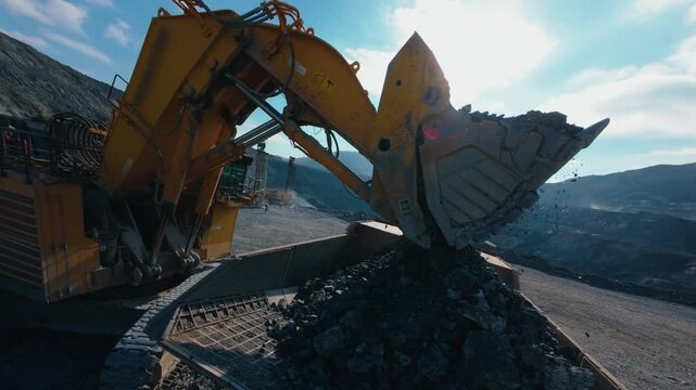 Tracking shot excavator loading gold ore to dump truck at open pit mine during daytime, aerial fpv.