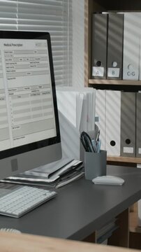 Vertical of desktop computer with medical form on screen standing on desk near window at reception in hospital, shelves with organized files visible in background