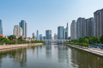 Fototapeta premium The Hai River flows through the heart of Tianjin, flanked by a mix of modern skyscrapers and green riverside promenades reflecting in the calm water. China, 22 May 2025