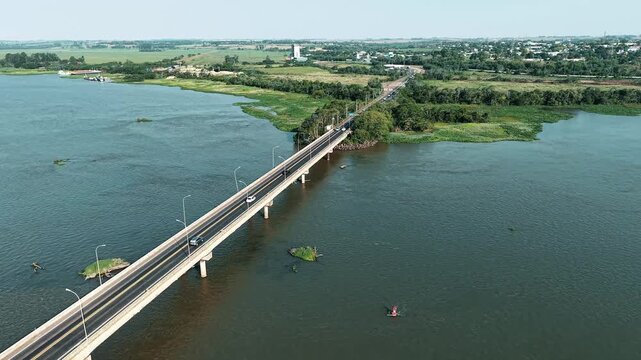 Aerial drone flyover of Ayrton Senna Bridge approaching Gua&iacute;ra, Brazil, with vehicles crossing Paran&aacute; River and entering the city via highway connection.