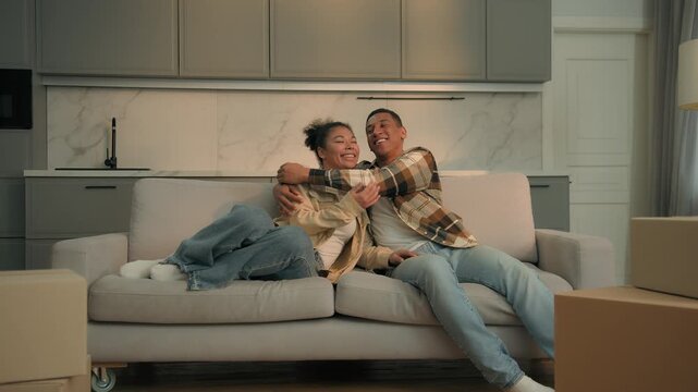 Smiling African American couple family unpack new couch after home renovation buying apartment moving day husband wife together celebrating sitting cuddling on sofa man woman house mortgage insurance 