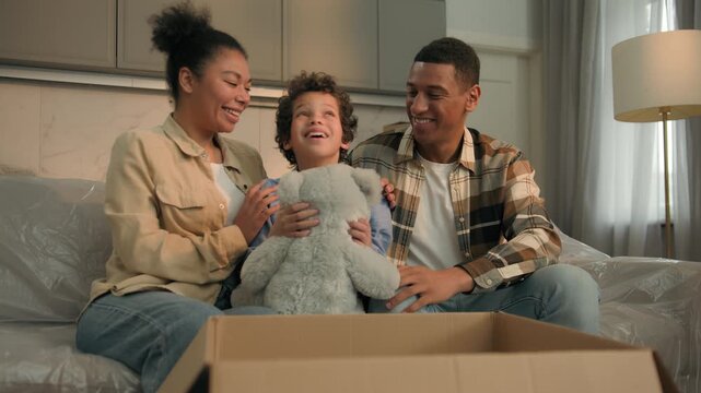 Happy African American family at home parents present giving gift to little kid boy child son open cardboard box parcel surprise birthday with toy teddy bear mother father unpacking stuff relocation