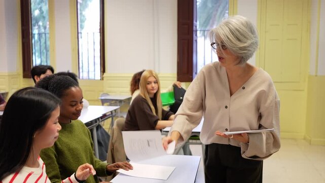 Senior female teacher walking among desks and distributing test sheets to a diverse group of college students before an exam or lesson