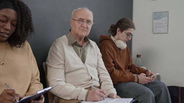 Medium portrait of senior Caucasian male patient sitting on chair with other people in hospital waiting room, looking at camera