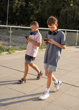 Two boys walking together in city while using smartphones, ignoring each other. Concept of digital isolation and reduced face to face communication in modern childhood.