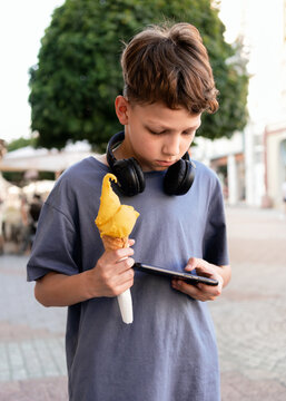 Boy holding yellow ice cream cone and using smartphone outdoors. Concept of Gen Alpha kids, mobile addiction, screen time, digital lifestyle, distraction during everyday activities.