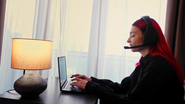 Woman at her workspace is providing virtual support while resolving technical issues efficiently. Lady at desk wearing headset is engaged in remote job session using laptop and talking to clients