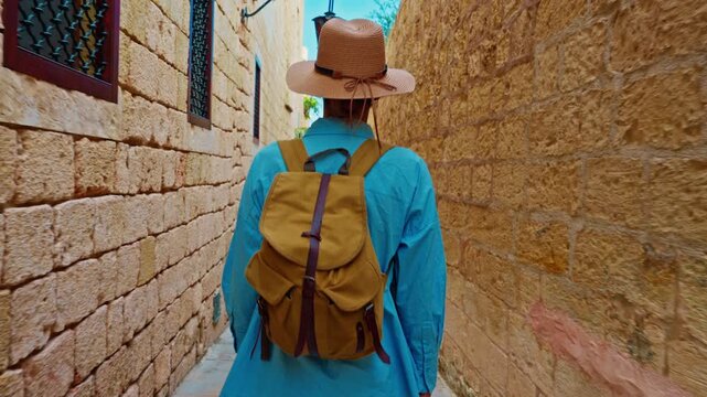 A female tourist in a hat and with a backpack walks along the traditional narrow old streets in Mdina, Malta