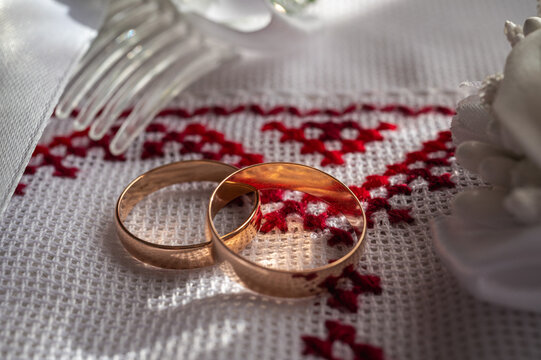 wo gold wedding rings on traditional Ukrainian embroidered rushnyk with white rose boutonniere and bridal hair accessory. Romantic marriage ceremony heritage concept