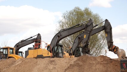 Rząd nowoczesnych koparek gąsienicowych stoi na placu budowy. Potężne, żółte maszyny z długimi ramionami są gotowe do robót ziemnych. W tle widać drzewa i błękitne niebo z delikatnymi chmurami © tomo81