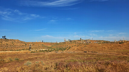Fototapeta premium Oil pumps across dry hillside landscape. Wide view of multiple oil pumpjacks operating across arid rolling hills under a bright blue sky in a remote energy field.