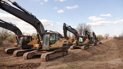 Rząd nowoczesnych koparek gąsienicowych stoi na placu budowy. Potężne, żółte maszyny z długimi ramionami są gotowe do robót ziemnych. W tle widać drzewa i błękitne niebo z delikatnymi chmurami © tomo81