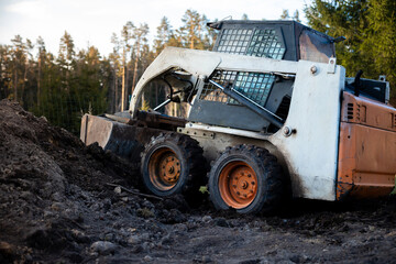 Fototapeta premium White and orange skid steer climbs a muddy slope, cab grid visible, thick tread tires caked with soil. Conifers surround cleared earth in daylight, low angle three quarter view.