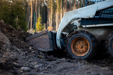 Fototapeta premium A skid steer loader with front bucket scoops soil at a forest edge site. Mud caked tires and hydraulic arms show wear in warm late day light by a wire fence.