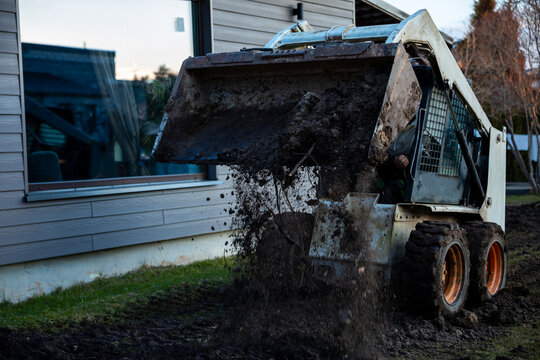 A skid steer with front bucket dumps wet soil by a modern house with gray siding. Orange rimmed tires are muddy, clods fly, motion blur shows late afternoon yard work.
