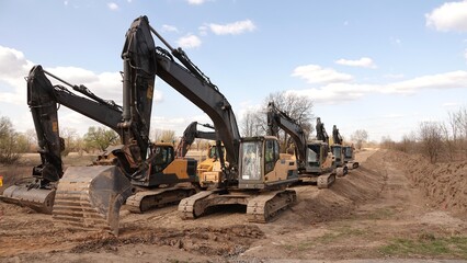 Rząd nowoczesnych koparek gąsienicowych stoi na placu budowy. Potężne, żółte maszyny z długimi ramionami są gotowe do robót ziemnych. W tle widać drzewa i błękitne niebo z delikatnymi chmurami © tomo81