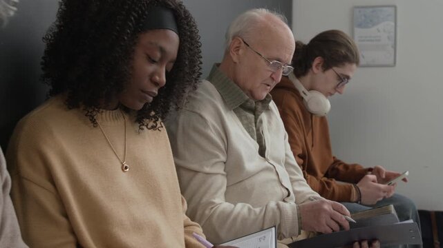 Group of diverse men and women sitting in hospital waiting room, signing documents, filling in forms, writing in notebook and using smartphone