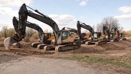 Rząd nowoczesnych koparek gąsienicowych stoi na placu budowy. Potężne, żółte maszyny z długimi ramionami są gotowe do robót ziemnych. W tle widać drzewa i błękitne niebo z delikatnymi chmurami © tomo81