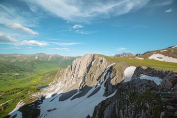 Scenic aerial dizzying top view to rocky cliff and sharp rocks under clouds in blue sky. Vertigo vivid alpine scenery with sheer crags above small lake in green dale. Colorful dizzy high mountains. © Daniil