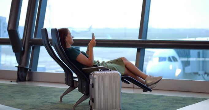 Female traveler waiting comfortably in airport lounge, sitting on reclined long chair and browsing smartphone while relaxing during international flight delay, slow motion. Airliner taxiing outside