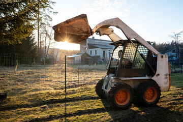 A compact skid steer with orange wheels lifts its bucket near a house under renovation. Late afternoon sun, long shadows, wire fence, muddy tracks, and weathered paint are visible. © True Pixel Art