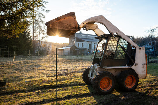 A compact skid steer with orange wheels lifts its bucket near a house under renovation. Late afternoon sun, long shadows, wire fence, muddy tracks, and weathered paint are visible.
