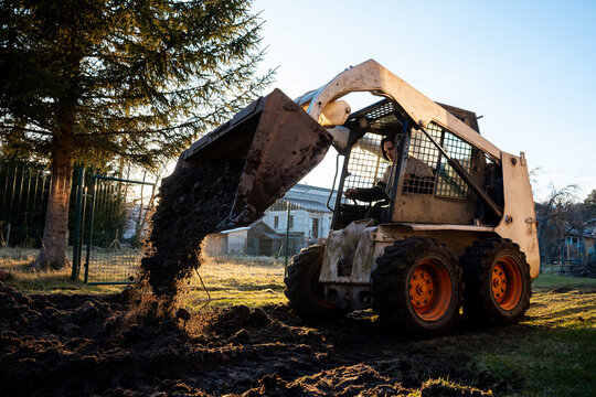 A skid steer with orange wheels tips its bucket, spilling dark soil beside a wire fence and conifer. Strong side light, flying clods, and low vantage emphasize motion.