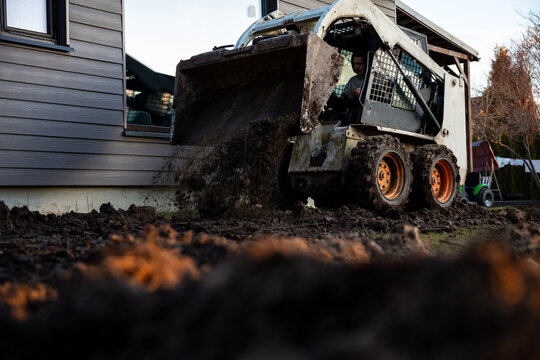 A compact skid steer dumps wet soil against a house foundation with gray siding. Late afternoon light highlights flying clods, muddy tires, ruts, and motion blur at ground level.