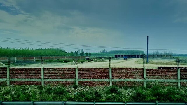 Long red-brick boundary walls enclose rural construction sites with partially built structures and open land, stretching alongside green fields and tree lines under a cloudy sky in India.