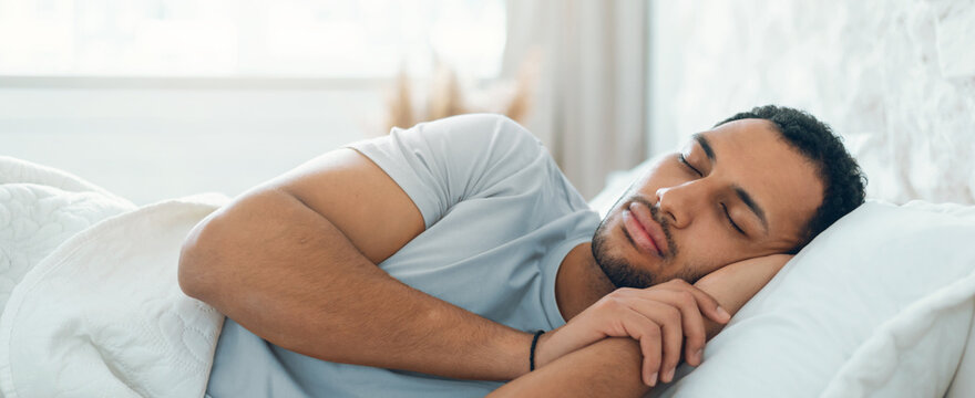 A man lies in bed with his eyes closed, resting on a pillow. Sunlight enters the room, creating a warm atmosphere. He appears comfortable and relaxed while sleeping in a cozy setting.