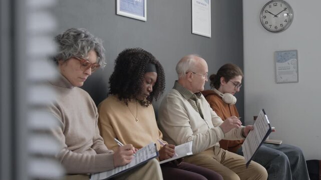 Medium long shot of group of diverse patients sitting on chairs in hospital hall, waiting in queue, checking time and making notes