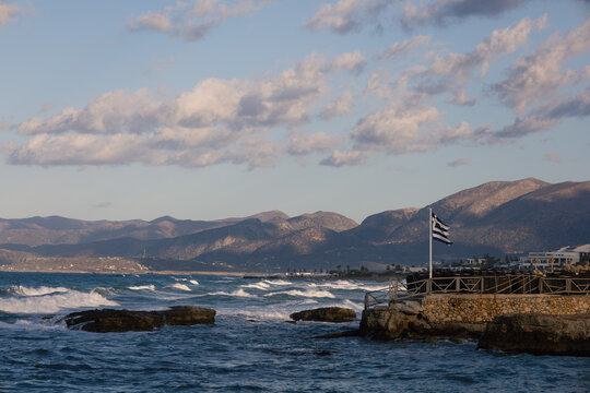 Scenic coastal landscape with rocky coast and mountains in Crete. Greek flag on a seaside pier under a soft cloudy sky. Travel concept. Waves of Aegean Sea. Amazing Hersonissos or Chersonissos