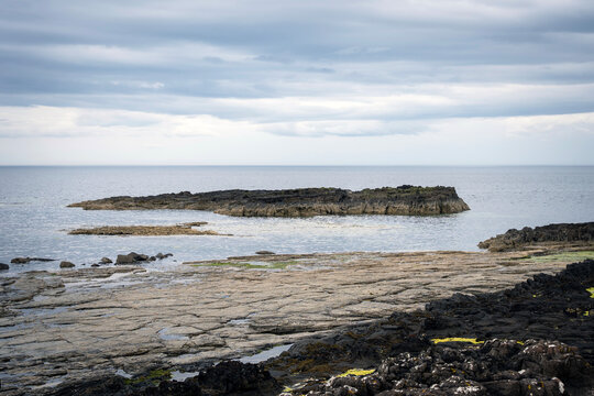 Dark volcanic rock formations protruding from the calm water under a cloudy sky on the coast of Scotland