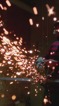 Man working at a factory cuts metal, vertical. Male at a construction site cuts off piece of metal, bright flying sparks and particles of material fly. Guy worker use angle grinder cutting metal