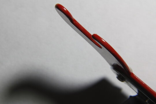 Fotograf&iacute;a macro de un abrelatas met&aacute;lico con mango rojo, tomada con poca profundidad de campo y fondo neutro. Iluminaci&oacute;n directa que resalta reflejos y texturas del metal.