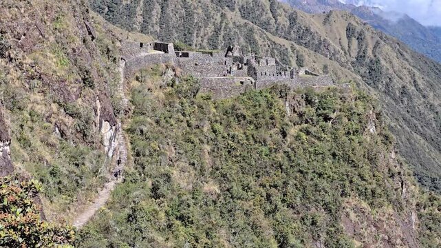 Cusco Region, Peru - 26 June 2024. Sayacmarca ruins perched on a high cliff along the Inca Trail, overlooking a deep green valley with steep slopes and distant mountains under light cloud.