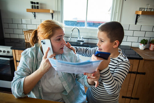 Stressed mother reviewing bills on phone while son seeks attention in home kitchen