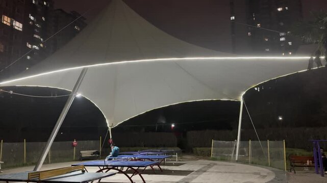 Nighttime scene of a public ping pong area in a residential neighborhood in China, illuminated by a modern canopy structure. People gather casually around outdoor table tennis tables, creating a relax