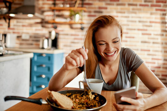 Smiling woman eating breakfast while using smartphone at home kitchen