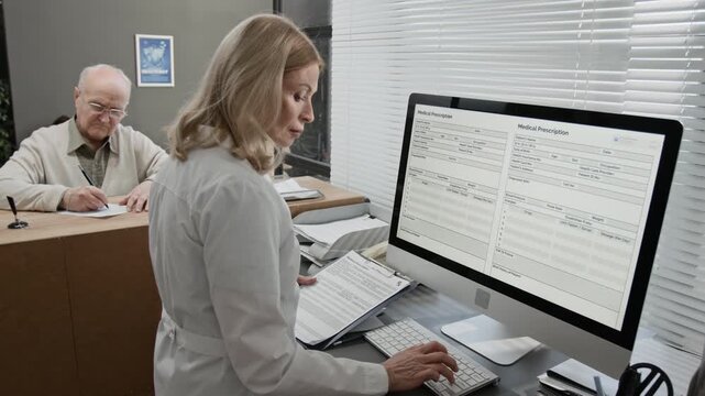 Medium shot of mature Caucasian female receptionist entering data on desktop computer at work in hospital, senior patient filling in form at desk in background