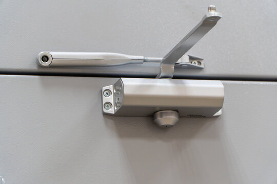 silver hydraulic automatic door closer mechanism with adjustable metal arm installed on top of gray fireproof entrance door in commercial building.