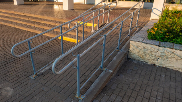 A concrete accessibility ramp featuring dual grey metal handrails leading to a building entrance, with stone-paved steps and a flower bed on the side under warm late afternoon sunlight.