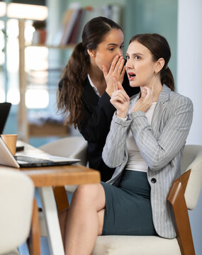 Curious female employee sharing gossip with surprised young colleague during workday in office