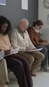 Vertical of diverse group of male and female patients sitting on chairs in clinic hall, waiting in queue, writing and scrolling on phone