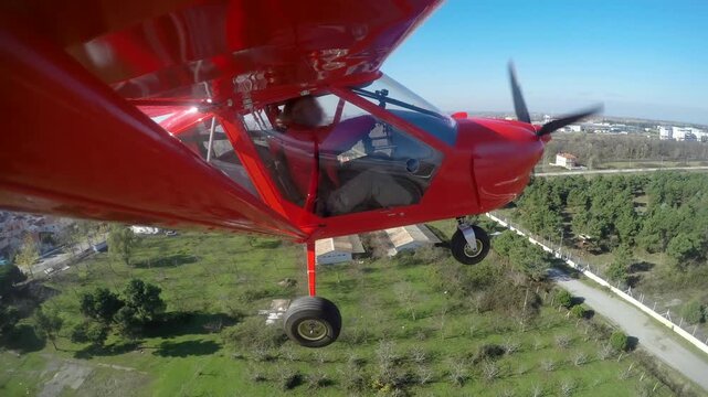 Red single seat propeller airplane approaches runway during controlled landing at airport. Small aircraft descends smoothly showing pilot cockpit view city below and spinning.