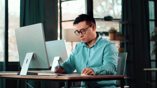 A man with glasses is focused on his work at a desk in a modern office. He writes in a notebook while using a computer. The workspace has large windows that let in natural light.
