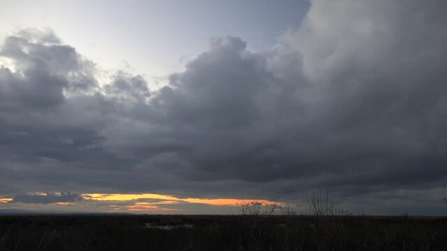 Sunset glowing behind dark nimbus clouds over flat plain landscape under dramatic sky. Evening glow reveals dusk light, approaching storm, and cloud formation plus cloud movement.