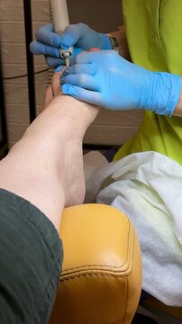 Close-up foot pedicure with electric file, podiatrist wearing blue gloves performs hardware debridement of thick fungal toenails in clinical salon, diabetic foot precautions observed, sterile.