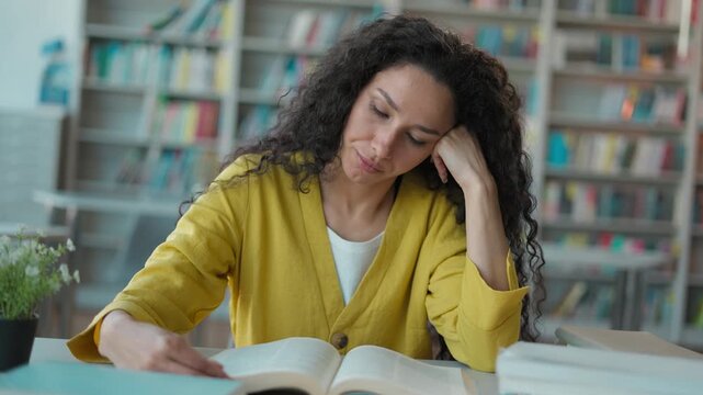 Tired Latino Arabian woman sitting reading book studying for exam in quiet library student girl female learning knowledge university college education lecture session desk boredom intelligence focus